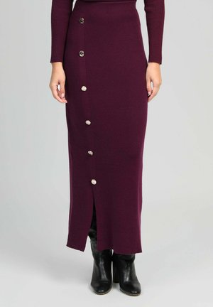 Jupe longue en tricot violette avec une fente sur le côté et des boutons décoratifs en argent sur le devant. Associée à des bottes noires à talons hauts.