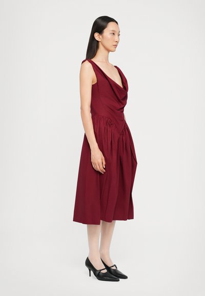 Burgundy draped dress with a fitted bodice, flared skirt, and gathering details; paired with black pointed heels.