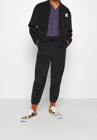 Chaqueta bomber negra con cremallera, camisa morada abotonada, pantalones jogger negros a juego y zapatillas coloridas con estampado y suelas blancas.