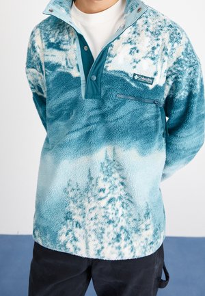 Persoon draagt een Columbia fleece trui met een blauwgroen en wit patroon, een ritszakje en een kraag met drukknopen.