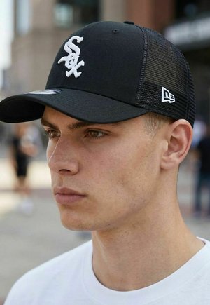 Junger Mann mit schwarzer Netz-Cap der Chicago White Sox und weißem Hemd, im Freien vor verschwommenem städtischem Hintergrund.