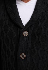 Cardigan nero a maglia con texture ondulata, caratterizzato da un colletto a scialle e due bottoni marroni, indossato sopra una maglietta bianca a scollo a V.