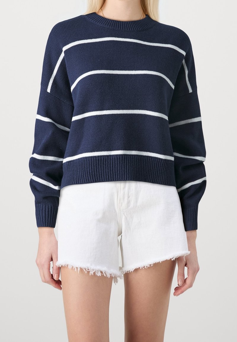 Femme portant un pull bleu marine avec des rayures blanches horizontales et un short blanc avec un ourlet effiloché, debout devant un fond uni.