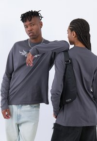 Le magliette grigie a maniche lunghe presentano una grafica sottile. Un modello indossa jeans chiari; l'altro ha una borsa a tracolla nera con un logo.