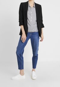 Schwarzes Blazer über einem grauweiß karierten Hemd, kombiniert mit hellblauen Jeans und weißen Sneakers. Slim Fit, lässiges Design, minimale Details.