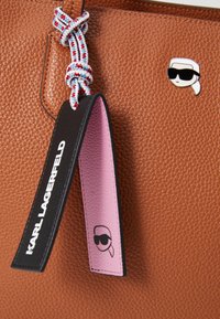 Sac en cuir texturé marron doté d'une sangle nouée multicolore, d'une étiquette noire et rose, et d'une épingle logo avec un design en silhouette.