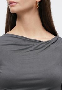 Γκρι τοπ με ντεκολτέ σε στυλ draped. Λείο ύφασμα, διακριτική λάμψη και μαλακή υφή. Χρυσό σκουλαρίκι σε σχήμα κρίκου προσθέτει μεταλλική νότα.