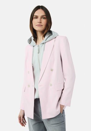 Helle rosa doppelte Blazerjacke mit zwei vorderen Taschen, über einem grauen Kapuzenpulli und grauen Hosen getragen. Glatte Stofftextur.