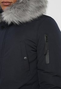 Person med en marinblå vinterjacka med en grå fuskpälskrage, knäppt ficka på bröstet och zip-ficka på ärmen.