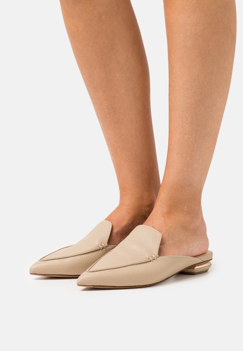 Jambes portant des ballerines beige en cuir à bout pointu, sans talon, à enfiler, avec des talons géométriques bas, sur un fond blanc.