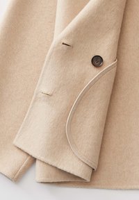 Beige wollen jas mouw met een enkele donkere knop, knoopsgaten en lichtgekleurde bies langs de rand.