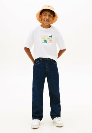 Garçon souriant portant un chapeau bob pêche, un T-shirt blanc à motif, un jean bleu foncé et des baskets blanches, debout les mains derrière le dos.
