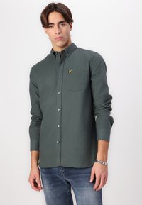 Cămașă button-up verde închis dintr-un material moale, având un buzunar pe piept și un mic logo galben, asortată cu blugi albaștri deschisi.
