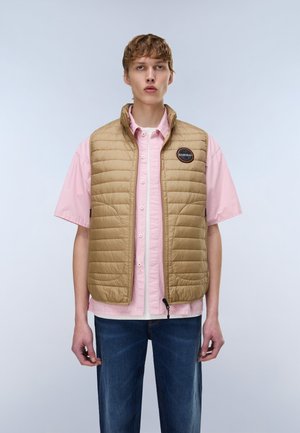 Ung mand iført en beige quiltet vest over en lyserød kortærmet skjorte med knapper og mørkeblå jeans, stående mod en ensfarvet baggrund.