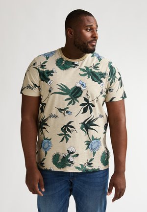 Camiseta beige de manga corta con estampado botánico en verde y azul, cuello redondo y corte cómodo, combinada con vaqueros azules.