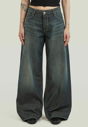 Personne portant un jean large délavé foncé et des chaussures noires, debout les bras détendus le long du corps, montrant le ventre et des tatouages sur les avant-bras.