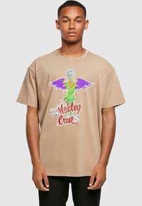 Beige kokvilnas T-krekls ar krāsainu, detalizētu pūķa grafiku ar purpura spārniem un tekstu "Motley Crue" sarkanā krāsā.