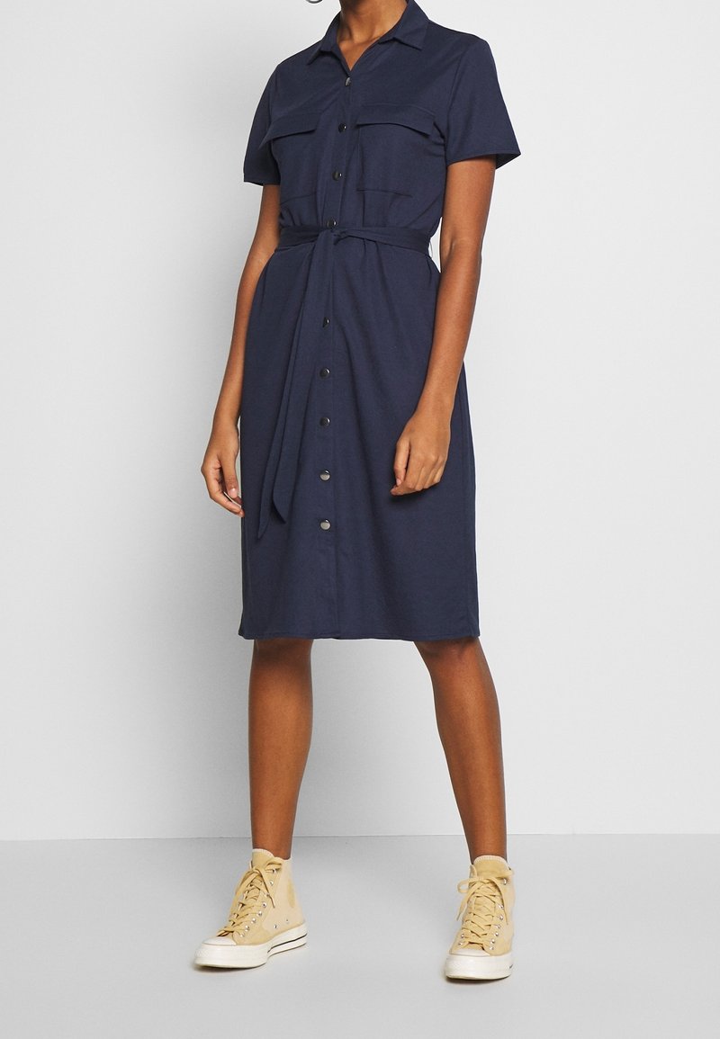 Personne portant une robe midi bleu marine à manches courtes boutonnée, avec poches poitrine et ceinture à la taille, associée à des baskets montantes beige.