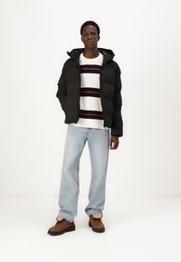 Sort pufferjakke med hætte, båret over en hvid sweater med vandrette røde og navy striber, parret med lyseblå jeans og brune støvler.