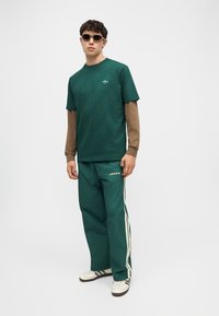 Άνδρας που φοράει πράσινο παντελόνι φόρμας Adidas, σκούρο πράσινο μπλουζάκι πάνω από μακρυμάνικο καφέ πουκάμισο, λευκά αθλητικά παπούτσια και γυαλιά ηλίου, στέκεται μπροστά σε λευκό φόντο.