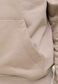 Sudadera de color beige claro con un bolsillo tipo canguro, puños y dobladillo acanalados. La tela parece suave y tiene una textura lisa.