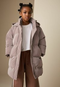 Világos taupe színű téli dzseki kapucnival és nagy, tömött szakaszokkal. Fehér, kivágott felső és barna melegítőnadrág fölött viselve.