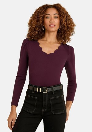 Burgundy hosszú ujjú bodysuit, fodros V-nyakkal, puha anyagból. Fekete, magas derekú farmerral és fekete övvel párosítva.