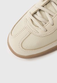 Béžová kožená teniska s texturovaným povrchem, kulatou špičkou, vícevrstvými švy, plochými tkaničkami a světle hnědou gumovou podrážkou.