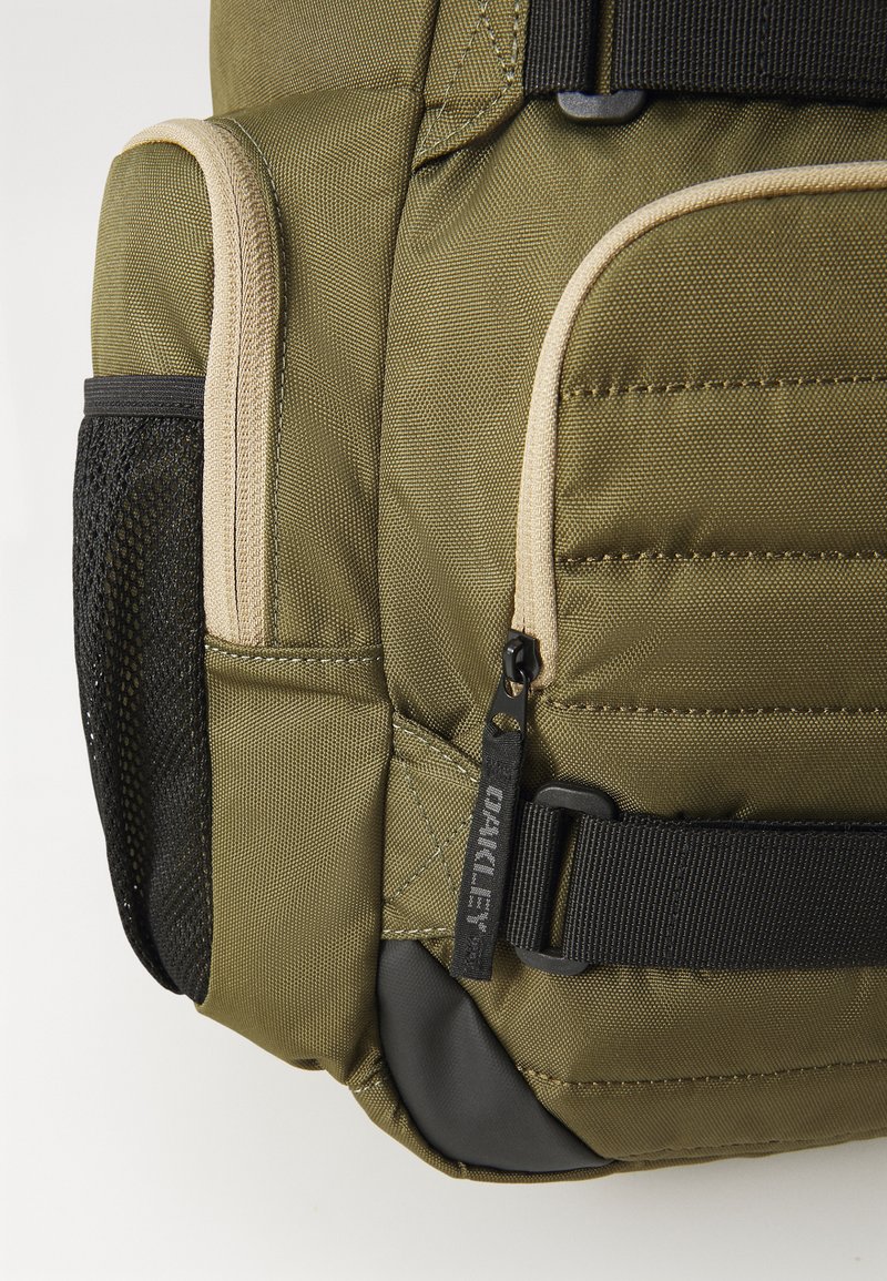 Zaino verde oliva con tessuto testurizzato, dettagli beige, tasca laterale in rete e un grande cursore nero con dettagli di branding.