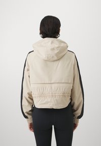 Afskaeret beige jakke med hætte, der har sorte sidepaneler, elastisk talje og glat tekstur. Bagfra ses den samlede detaljering.