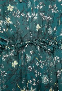 Vestido floral con fondo verde azulado, con flores en tonos verde claro y blanco. Cintura con volantes y detalle de lazo. Tejido suave y ligero.