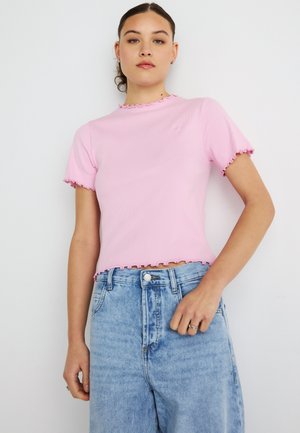 Tricou roz scurt, din material reiat, cu mâneci scurte și margini cu volănașe, asortat cu pantaloni scurți din denim albastru deschis, talie înaltă.