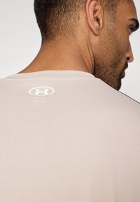 Lys beige treningsskjorte med en jevn tekstur, med en hvit logo på bakre nakke og svarte detaljer langs skuldrene.