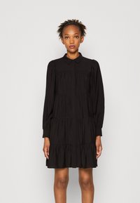 Svart lagerklänning i lätt tyg. Har långa ärmar, spetskrage och knappstängning fram. Lätt flödande silhuett.