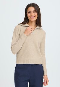 Pull en maille beige à côtes avec un col montant zippé et des manches longues, fabriqué en matériau tricoté texturé. Associé à un pantalon bleu marine.