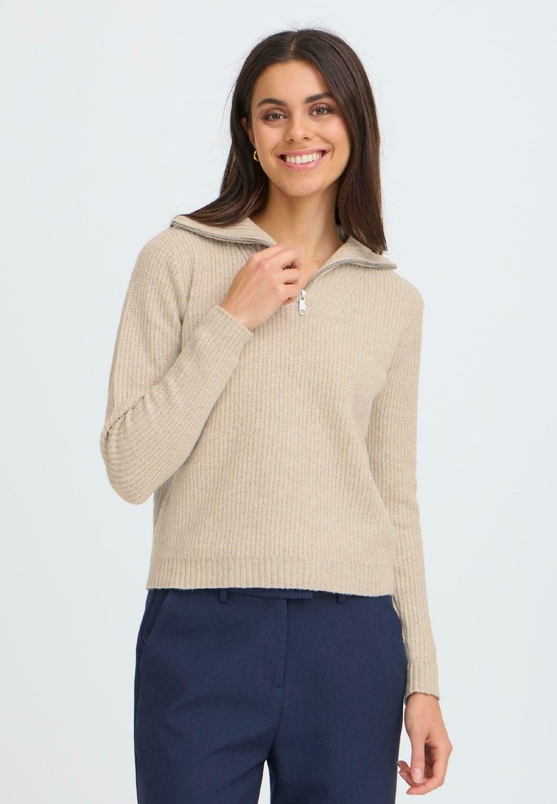 Pull en maille beige à côtes avec un col montant zippé et des manches longues, fabriqué en matériau tricoté texturé. Associé à un pantalon bleu marine.