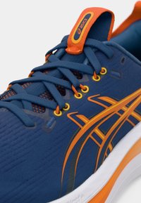 Scarpa da corsa con tomaia in mesh blu, accentata da dettagli arancioni, modelli testurizzati e suola bianca. Presenta occhielli gialli e lacci piatti.