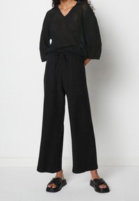 Zwarte gebreide top met een V-hals en korte mouwen, gecombineerd met zwarte wijde broek. Zwarte platte sandalen maken de outfit compleet.