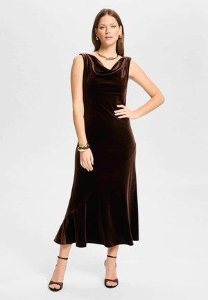 Femme portant une robe midi sans manches en velours marron foncé avec un décolleté drapé, associée à des talons noirs à bride à la cheville et des bijoux dorés.