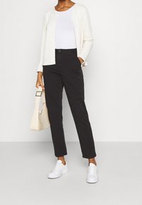 Weißer, gesteppter Cardigan, weißes Oberteil, schwarze Hose und weiße Turnschuhe. Die Handtasche ist hellbeige mit Krokodilmuster.