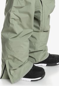 Pantalons de neige vert clair, résistants à l'eau, avec des poignets zippés, associés à des chaussures de sport noires avec des semelles blanches et un matériau texturé.