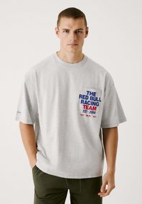 Graues Baumwoll-T-Shirt mit kurzen Ärmeln, das einen blau-roten Grafikdruck mit der Aufschrift "DAS RED BULL RACING TEAM GEGRÜNDET 2004" auf der Brusttasche zeigt.
