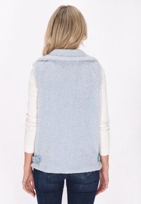 Helles blaues Fleece-Weste mit weicher, texturiertem Oberfläche, mit einem Kragen und Knöpfen an den Seiten. Über einem weißen Langarmshirt getragen.