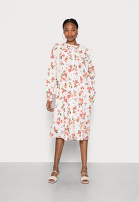 Vestido floral com base branca, apresentando flores vermelhas e azuis, gola com babados e mangas longas. Combinado com sandálias rasas de cor clara.