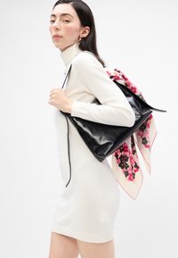 Geantă de mână din piele neagră cu un finisaj neted, având o eșarfă din mătase cu model floral legată ca un accent, expusă pe un fundal deschis.