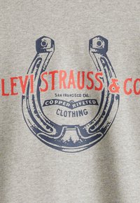 Szürke kapucnis pulóver, nyomtatott tengerészkék patkó grafikával és piros felirattal: "LEVI STRAUSS & CO", további márkázási részletekkel alatta.