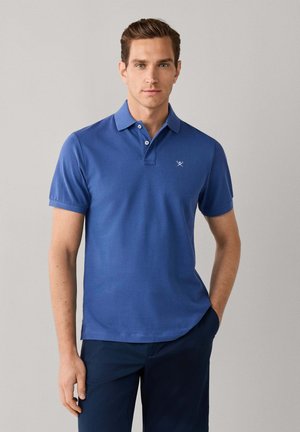 Jeune homme aux cheveux courts et bruns, portant un polo bleu à manches courtes et un pantalon foncé, debout avec une main dans la poche sur un fond uni.