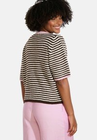 Raibi adīšanas džemperis brūnā un krēmkrāsas krāsā, ar īsām piedurknēm un rozā apdari. Modelis valkā gaiši rozā bikses, demonstrējot džempera aizmuguri.