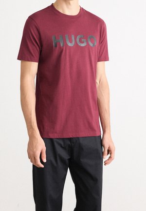 Homme portant un T-shirt bordeaux à manches courtes avec "HUGO" imprimé en grandes lettres noires, assorti à un pantalon noir.