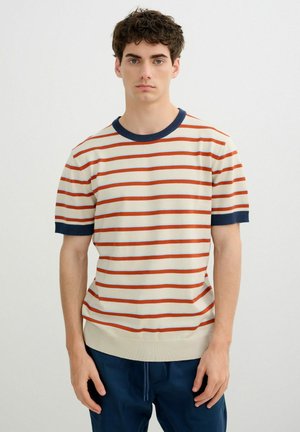 Joven con el pelo corto y rizado que lleva una camisa de manga corta color crema con rayas naranjas y ribetes azul marino, combinada con pantalones azul marino.